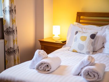 A bed with rolled white towels on top next to a wooden nightstand with a lamp and a pillow with leaf patterns at Harbour Lights House in Seahouses