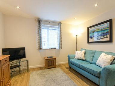 A living room with a teal sofa and white cushions a floor lamp a framed beach painting a window with curtains a small wooden table a tv on a glass stand and wooden cabinet at Braidcarr Cottage in Seahouses