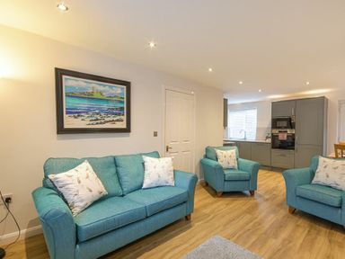 A living room with blue sofas and armchairs and a kitchen area with grey cabinets in the background at Braidcarr Cottage Seahouses
