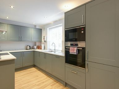 A kitchen with grey cabinets wooden floor built in ovens and a window at Braidcarr Cottage in Seahouses