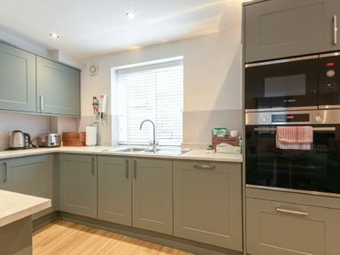 A kitchen with grey cabinets a window above a sink a kettle a toaster and built in oven at Braidcarr Cottage in Seahouses
