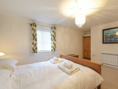 A bedroom with a double bed white pillows towels on bed window with curtains wooden wardrobe and framed painting on wall at Braidcarr Cottage in Seahouses