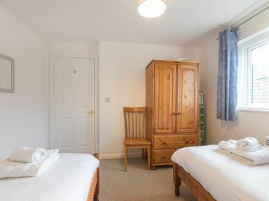 A bedroom with two single beds with white linens a wooden wardrobe and chair and a window with blue curtains at Braidcarr Cottage in Seahouses