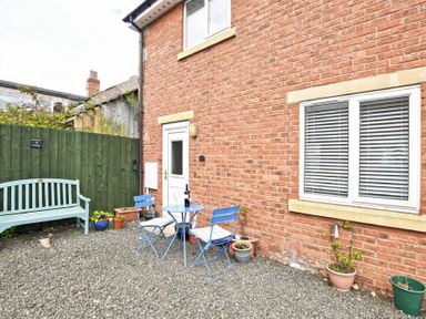 A small outdoor patio with a green wooden bench blue table and chairs and potted plants next to a brick house at Braidcarr Cottage in Seahouses