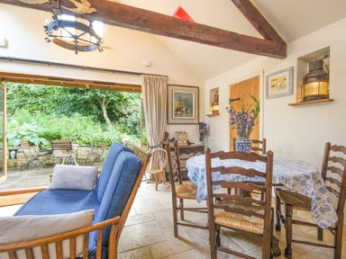 A dining area with wooden chairs and a table with a vase of flowers and a blue sofa next to large open doors showing a garden at The Byre at Warkworth