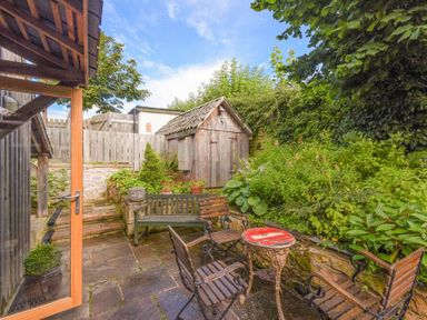An outdoor patio area with wooden chairs a small round table a wooden bench stone steps greenery and a small wooden shed at The Byre at Warkworth in Warkworth