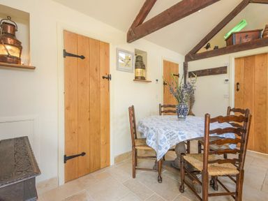 A dining area with a round table covered with a tablecloth and four wooden chairs in a room with wooden doors and exposed ceiling beams at The Byre at Warkworth
