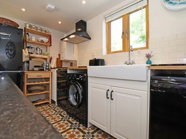 A kitchen with a black refrigerator a wooden shelf a washing machine a white sink under a window and patterned floor tiles at The Byre at Warkworth in Warkworth