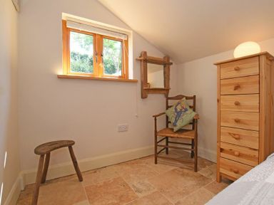 A small room with a wooden chair with a cushion a wooden stool a dresser with drawers and a window at The Byre at Warkworth in Warkworth
