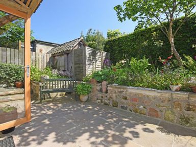 A garden patio with stone tiles a wooden bench flower pots and a small wooden shed surrounded by plants at The Byre at Warkworth