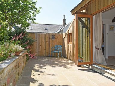 A patio area with a wooden fence a blue chair plants and an open glass door leading indoors at The Byre at Warkworth in Warkworth