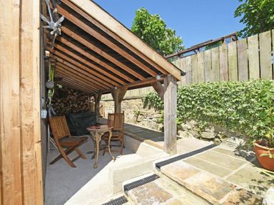 A covered outdoor seating area with wooden chairs and a small table next to a stone wall and plants at The Byre at Warkworth in Warkworth