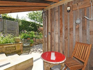 A covered outdoor seating area with a wooden chair and a round red table near a garden with plants at The Byre at Warkworth