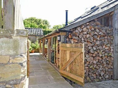A woodpile stacked against a building with a wooden gate and stone pathway at The Byre at Warkworth
