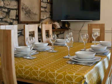 A dining table with plates bowls and wine glasses set on a yellow patterned tablecloth in a room with a stone wall and wooden chairs at Lobster Lodge in Seahouses