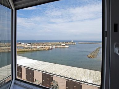 A view of a harbor with boats docked and a breakwater seen through an open window at Lobster Lodge in Seahouses