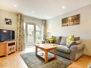 A living room with a grey sofa, wooden coffee table, television on a wooden stand and glass door with floral curtains at East Havers in Alnwick