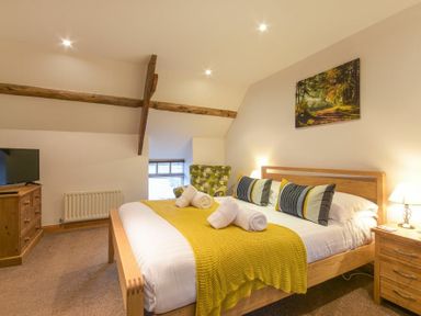 A bedroom with a wooden bed with yellow throw and rolled towels a wooden dresser with a tv and a green patterned armchair at East Havers in Alnwick