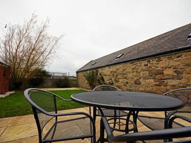 An outdoor patio with metal table and chairs on a stone floor next to a grassy area and a stone building at East Havers in Alnwick