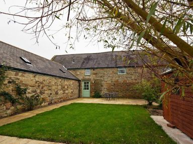 A backyard with green grass stone walls a door a small table with chairs and a wooden shed at East Havers in Alnwick