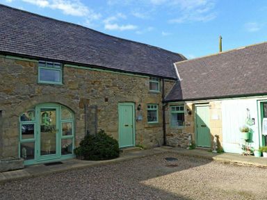 An exterior view of stone buildings with green doors and windows surrounding a gravel courtyard at East Havers in Alnwick