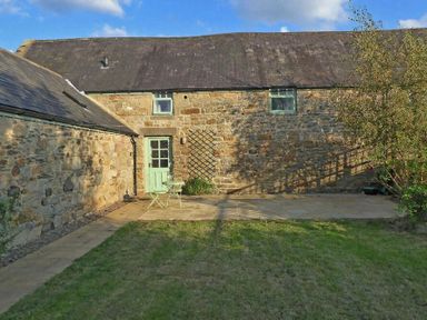 A stone building with a green door and two small green windows a paved patio with a small table and two chairs and a grassy lawn with a tree at East Havers in Alnwick