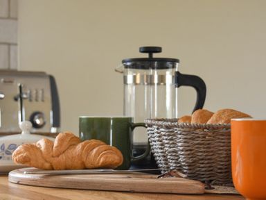 A kitchen counter with a croissant on a wooden board a green mug a glass French press a basket of croissants and an orange mug at Rock Lodge in Alnmouth