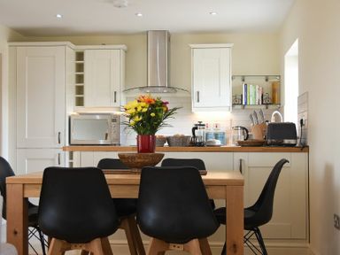 A kitchen with white cabinets and a wooden dining table with black chairs and flowers on the table at Rock Lodge in Alnmouth