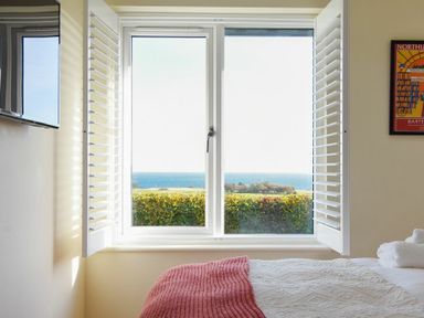 A bedroom window with white shutters overlooking a garden and sea with part of a bed visible at Rock Lodge Alnmouth