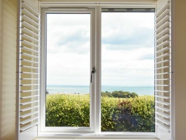 A window with white shutters looking out onto greenery and the sea at Rock Lodge in Alnmouth