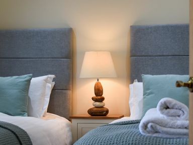A bedroom with two beds featuring grey headboards a bedside table with a lamp and folded towels at Rock Lodge in Alnmouth