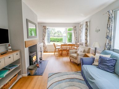 A living room with a fireplace a blue sofa a beige armchair a wooden dining table and windows with floral curtains at Manor Cottage in Embleton