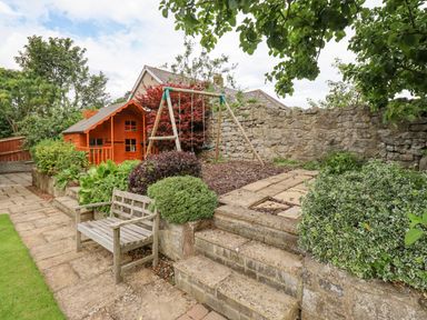 A garden with a wooden bench a children's swing set a wooden playhouse stone steps and a stone wall at Manor Cottage in Embleton