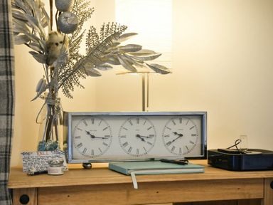 A wooden table with a clock showing three time zones a glass vase with decorative branches a notebook and a black device with a dock at West End Cottage in Embleton