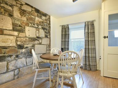 A dining room with a round wooden table and four chairs next to a stone wall and window with plaid curtains at West End Cottage in Embleton