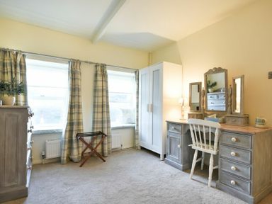 A bedroom with two windows covered by patterned curtains a white wardrobe a dresser with a plant a vanity table with a chair and mirrors at West End Cottage in Embleton