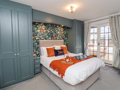 A bedroom with a bed, floral wallpaper behind the bed, built-in wardrobes, and glass doors leading to a conservatory at The Bolthole (Seahouses) in Seahouses