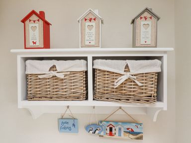 A white wall shelf with two wicker baskets and three decorative house-shaped plaques on top at The Bolthole (Seahouses) in Seahouses
