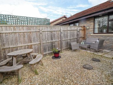 A fenced outdoor patio with gravel ground a wooden picnic table a potted plant and a seating area with wicker furniture at The Bolthole (Seahouses) in Seahouses