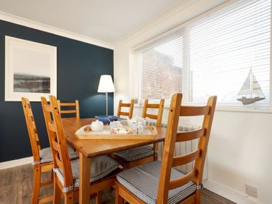 A dining room with wooden table and chairs next to a window with blinds at Stones Throw in Beadnell