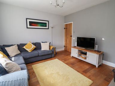 A living room with a gray sofa, yellow and beige cushions, a yellow rug, a wooden TV stand with a TV, a wooden door, and a floor lamp at Alnside Cottage in Alnmouth