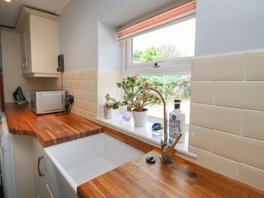 A kitchen countertop with a white sink and faucet next to a window with plants and bottles on the windowsill at Alnside Cottage in Alnmouth