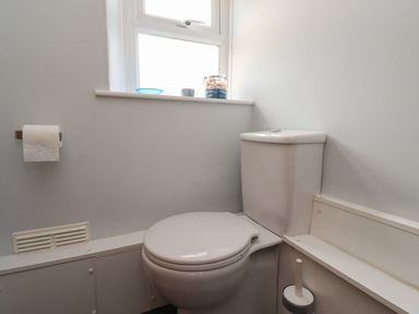 A bathroom with a toilet roll holder, closed toilet seat, brush holder, and window with small items on the sill at Alnside Cottage in Alnmouth
