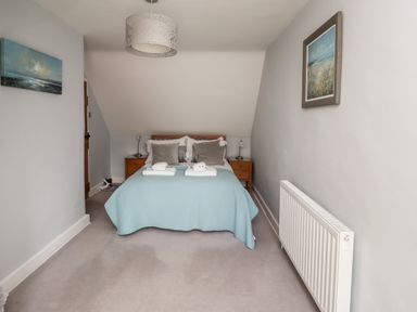 A bedroom with a double bed covered in a blue bedspread two bedside tables with lamps paintings on walls and a radiator at Alnside Cottage in Alnmouth