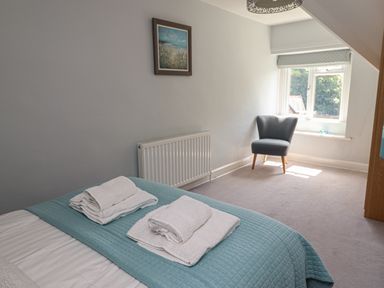 A bedroom with a bed covered by a blue quilt and towels on top a chair near a window and a framed picture on the wall at Alnside Cottage in Alnmouth