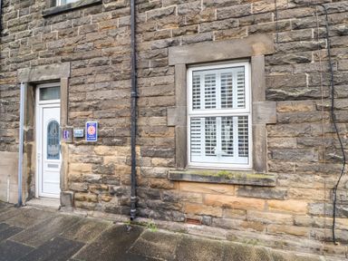 The exterior wall of a stone building with a white door numbered 17 and a window with white shutters and a small bird figurine on the window sill at Number Seventeen in Amble