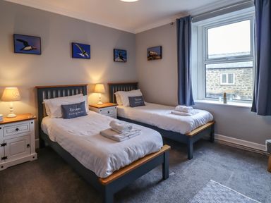 A bedroom with two single beds with white bedding blue cushions lamps on bedside tables and a window with blue curtains at Number Seventeen in Amble