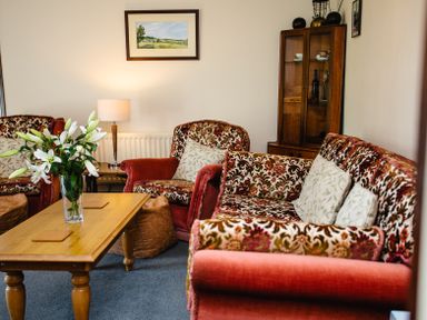 A living room with floral patterned armchairs and sofa a wooden coffee table with flowers a lamp and a display cabinet at Swallows Cottage nr Warkworth in Acklington