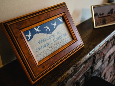 A framed embroidery with the text Welcome to Swallows Cottage on a wooden mantelpiece with another framed photo at Swallows Cottage nr Warkworth in Acklington