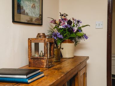 A wooden sideboard with books a rustic lantern a brass container and a vase with flowers at Swallows Cottage nr Warkworth in Acklington